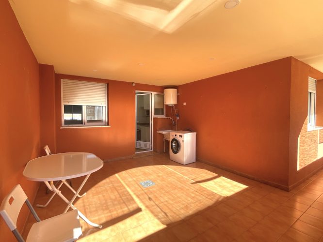 Primer plano de la terraza desde el que se ve una mesa, dos sillas, el acceso a la zoina, una ventana que da al interior de la vivienda, una lavadora y un calentador eléctrico