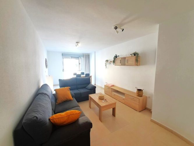 Primer plano del salón con un sofá grande y otro pequeño, una mesa pequeña y otra grande, una lámpara de pie, cuatro sillas, un mueble para televisión, un pequeño armario, un estante, un reloj de pared y un ventanal.