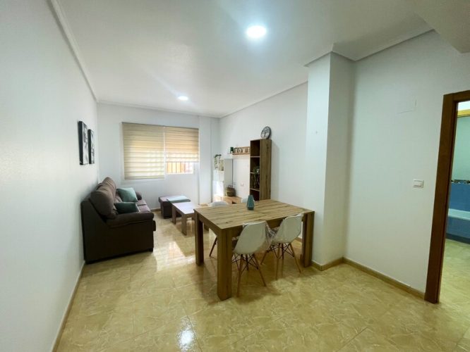 Primer plano del salón con una mesa y cuatro sillas, una mesa pequeña, un sofá, dos estanterías, un mueble para televisión, una ventana y dos estantes