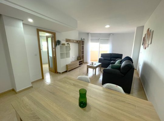 Segundo plano del salón con una mesa grande con cuatro sillas, dos sofás, una mesa pequeña, dos estanterías, un mueble para televisión, un estante, acceso a la cocina y un ventanal que da al balcón