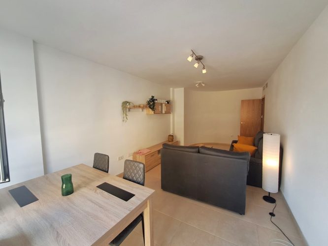 Segundo plano del salón desde el que se ve una mesa con 4 sillas, dos sofás, una mesita de noche, un mueble para televisión, un estante y una lámpara