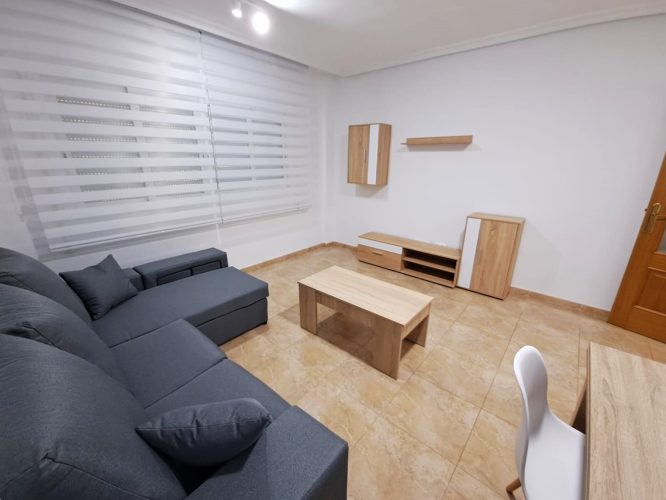 Segundo plano del salón con un sofá en forma de L, una silla, una mesita, un mueble para televisión, una ventana, un armarito de pared, un estante y un armarito en el suelo