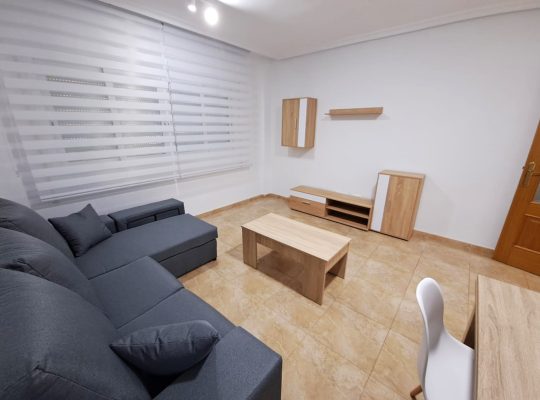 Segundo plano del salón con un sofá en forma de L, una silla, una mesita, un mueble para televisión, una ventana, un armarito de pared, un estante y un armarito en el suelo